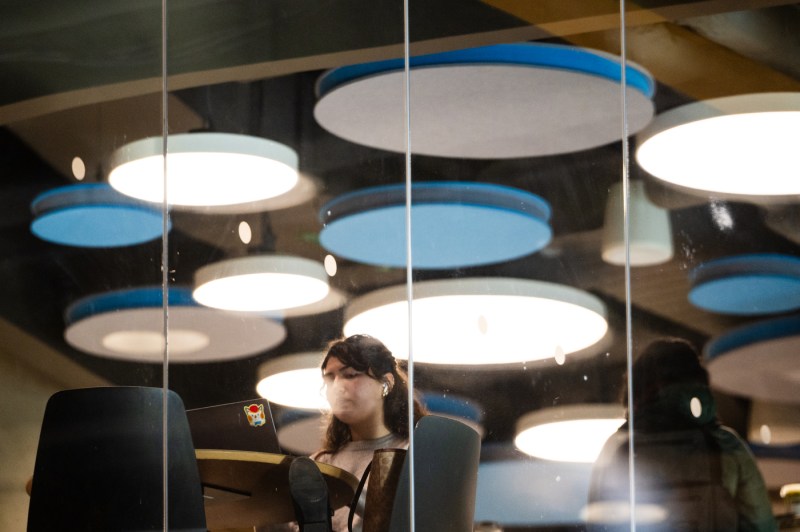 Photographed through a glass wall, students study at a table. Large, circular lights hang above. 