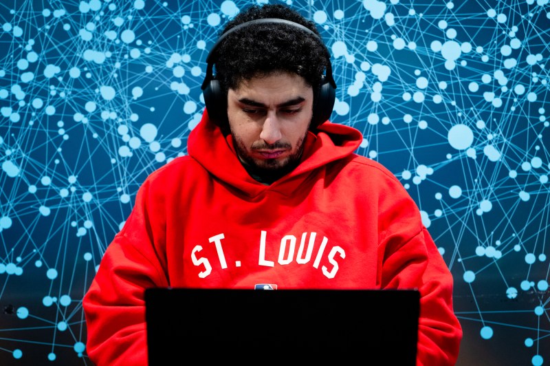 A student wearing a hoodie with the text "St. Louis" and headphones works on a laptop.