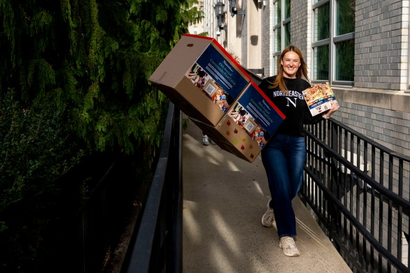 A person carries two boxes in one hand and food products in the other hand. The person is walking outside and is smiling.