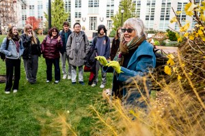 A group of Northeastern students stand outside in the grass on campus while listening to a woman speak.