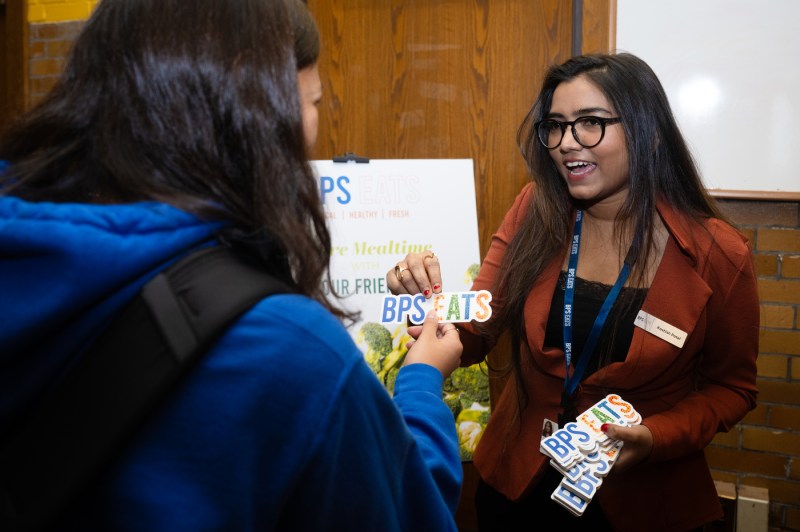 Two women in conversation, one wearing a blue hoodie shown from behind, and another wearing glasses, an orange blazer, and a name tag, giving out stickers.