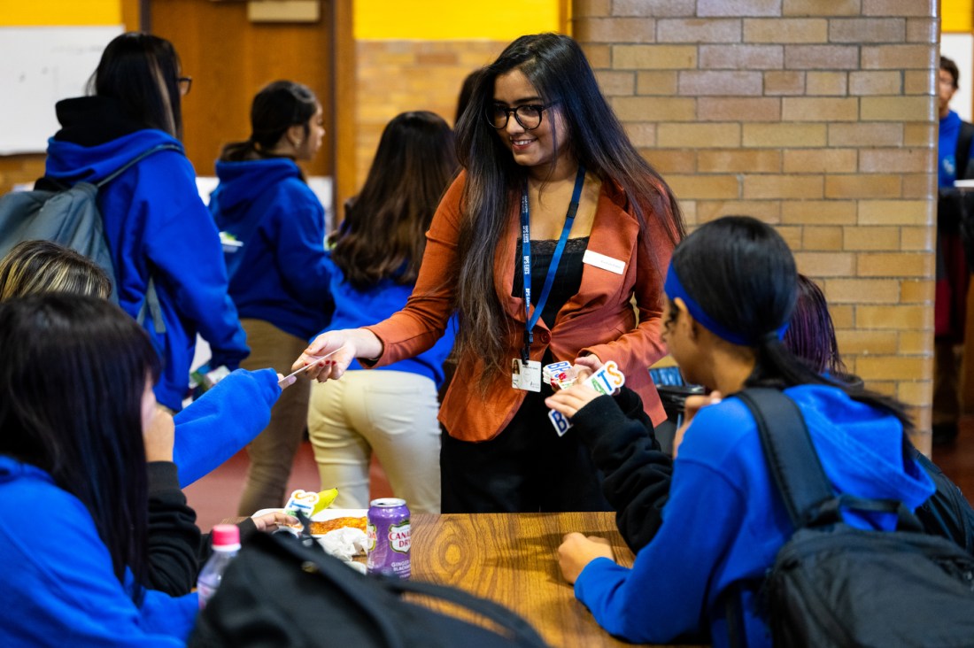 A woman with long dark hair and glasses, wearing an orange and white striped shirt and lanyard, stands at a wooden table distributing materials to students in blue hoodies who are seated around the table in a school cafeteria.