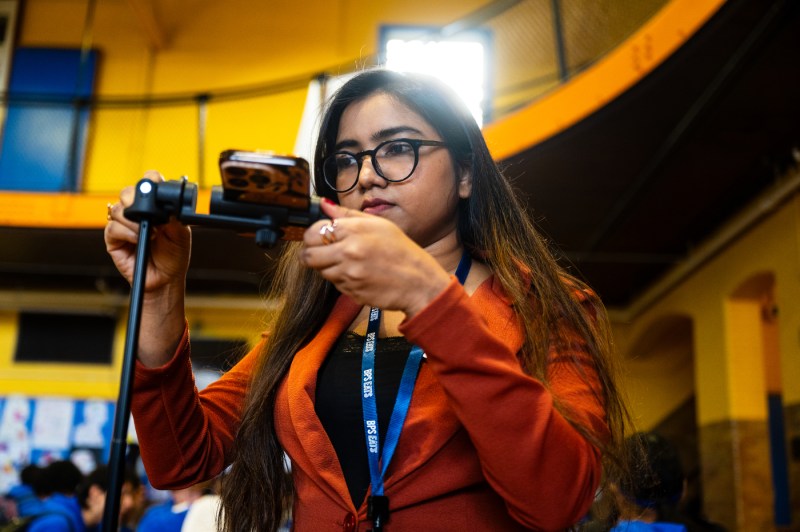 A young woman with long dark hair, glasses, and wearing an orange blazer with a blue lanyard takes a photo with her smartphone in a cafeteria with yellow walls.