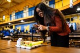 A young woman with long dark hair and glasses stands in a cafeteria, taking a photo of her meal tray with her smartphone. The tray contains a sandwich, banana, lettuce, and a carton of milk.
