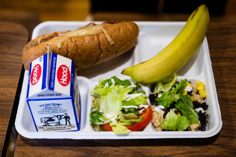 A cafeteria meal tray containing a sandwich, a banana, a small carton of milk, and a fresh garden salad with lettuce, tomatoes, corn, and black beans on a white rectangular plate.