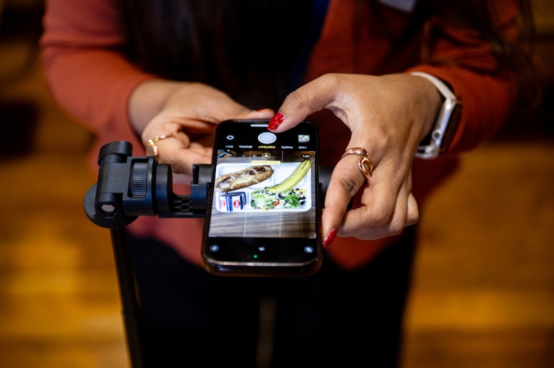 Close-up of hands holding a smartphone with a camera grip attachment, displaying a photo of a cafeteria meal tray with a sandwich, banana, milk carton, and salad on the phone's screen.