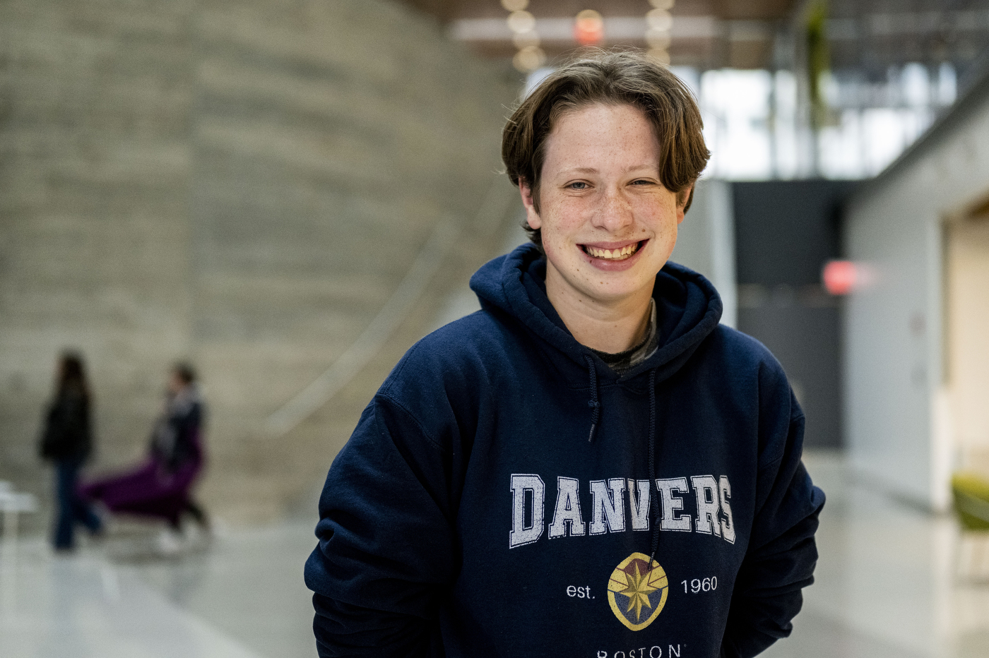 A smiling young person wearing a navy blue Danvers hoodie stands in what appears to be an indoor institutional setting with stairs and people visible in the blurred background.