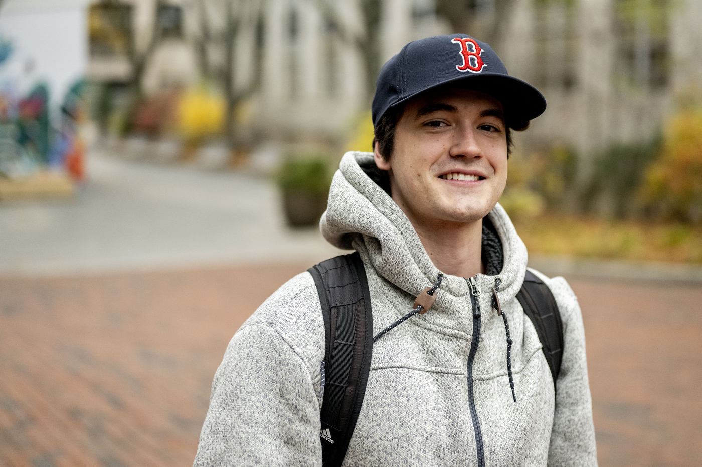 A smiling young man wearing a gray hoodie, backpack, and Boston Red Sox baseball cap stands outdoors on a brick plaza with trees and buildings visible in the background.