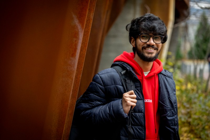 A person holding their backpack, which is on one shoulder, smiles for a photo outside.