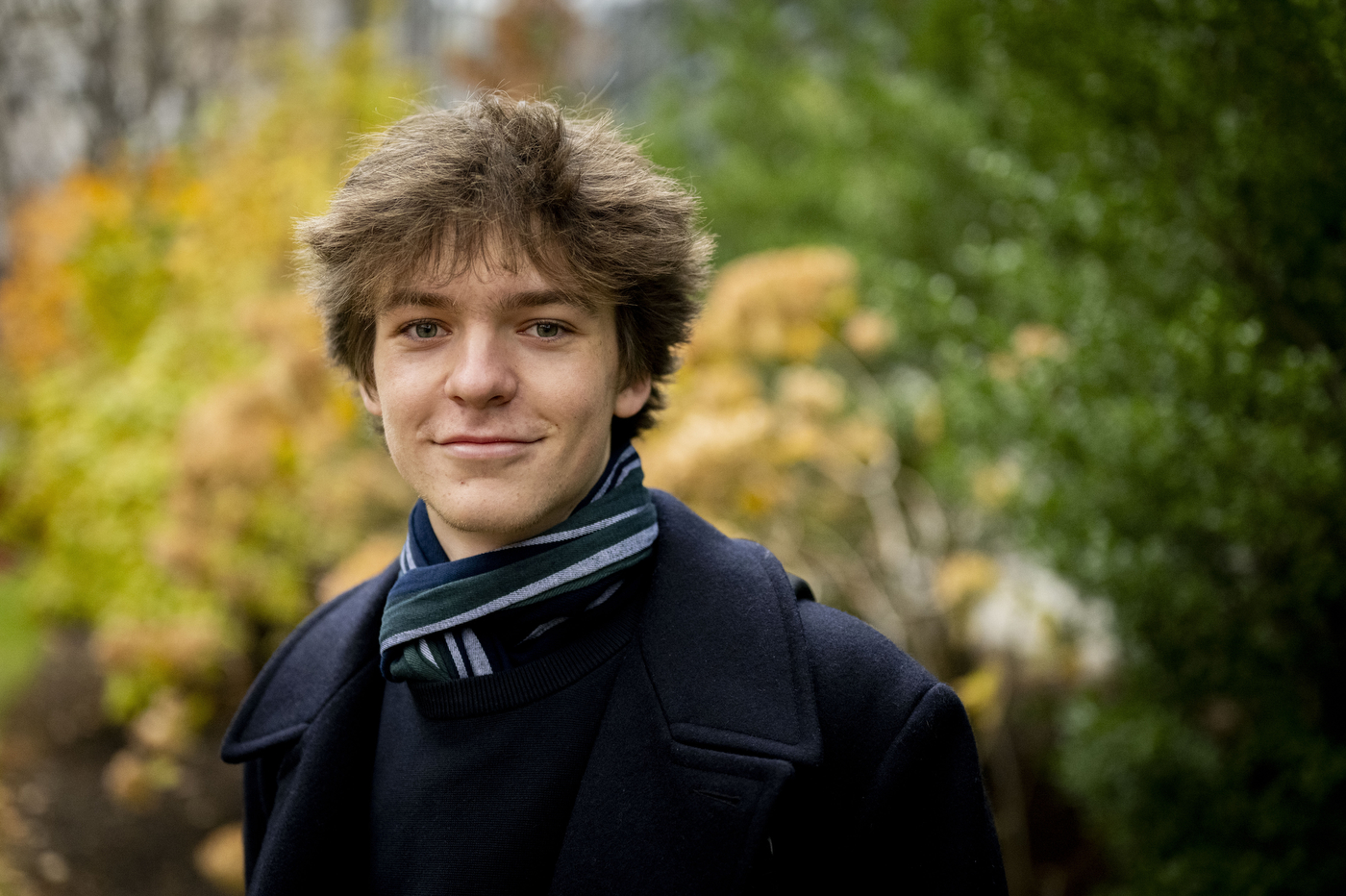 A young man with wavy brown hair wearing a dark coat and striped scarf stands outdoors with colorful fall foliage and greenery in the soft-focus background.