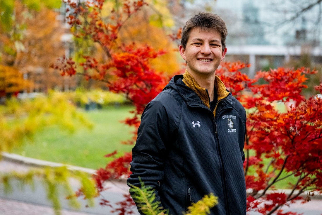 A smiling young man wearing a black jacket stands outdoors surrounded by vibrant red and yellow fall foliage, with buildings visible in the blurred background.