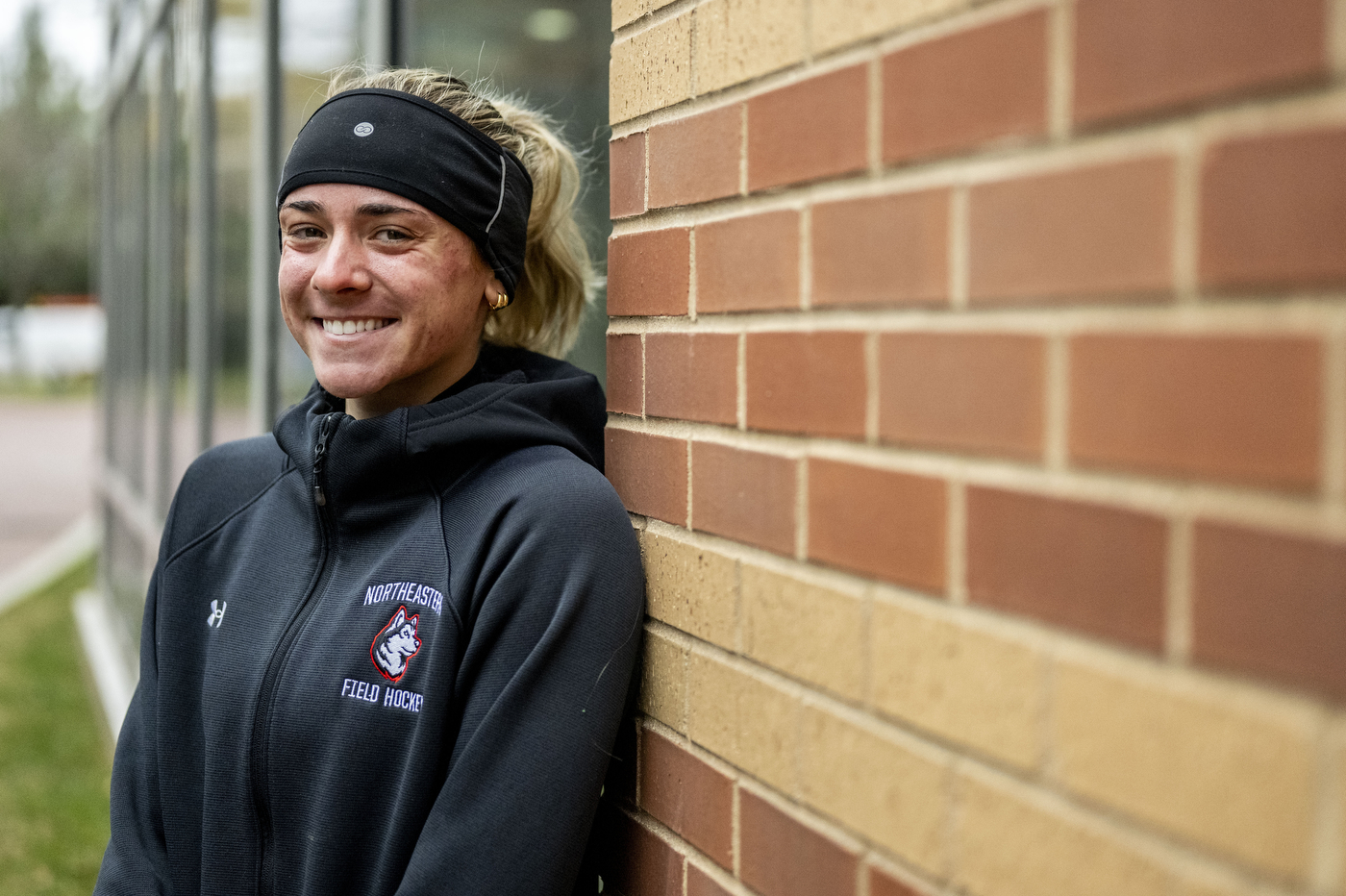 A smiling young woman wearing a black headband and Northeastern field hockey jacket leans against a brick wall outdoors.