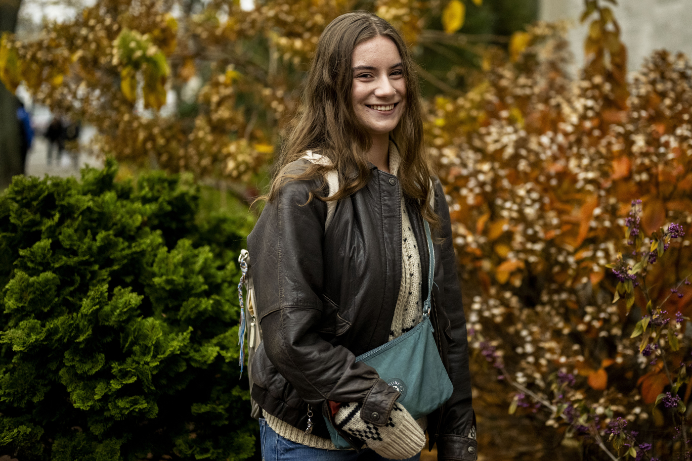 A smiling young woman with long brown hair wearing a black leather jacket, beige knit sweater, and teal crossbody bag stands outdoors with evergreen shrubs and golden fall foliage in the background.
