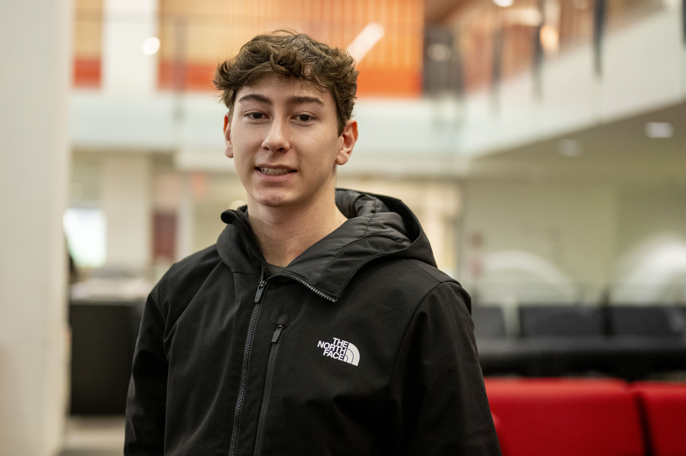 A young man with curly brown hair wearing a black North Face jacket stands inside the Curry Student Center at Northeastern University with red seating and modern interior architecture visible in the background.