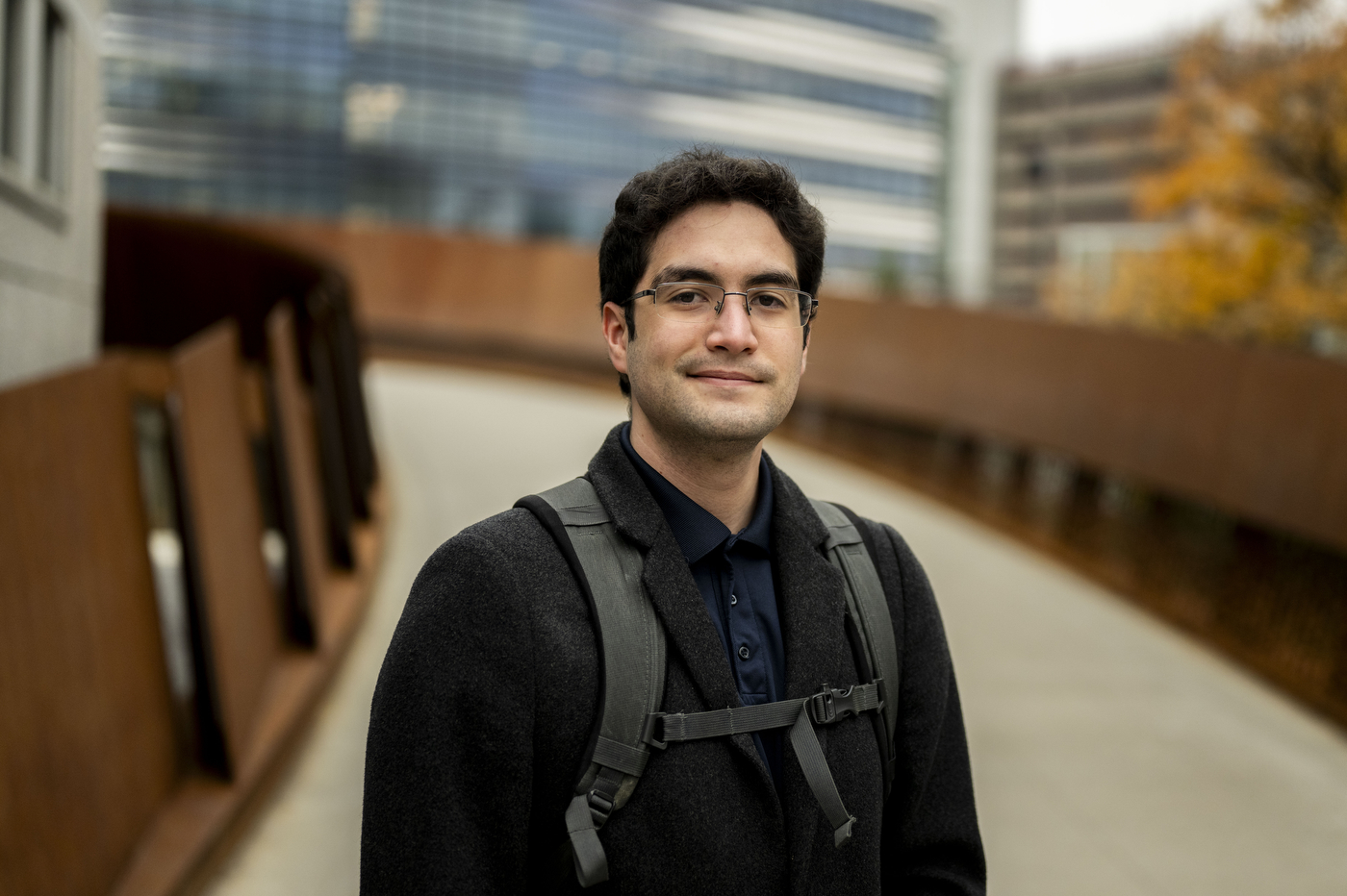 A young man wearing glasses, a dark button-up shirt, and a backpack stands on a modern campus walkway with contemporary buildings and fall foliage visible in the background.