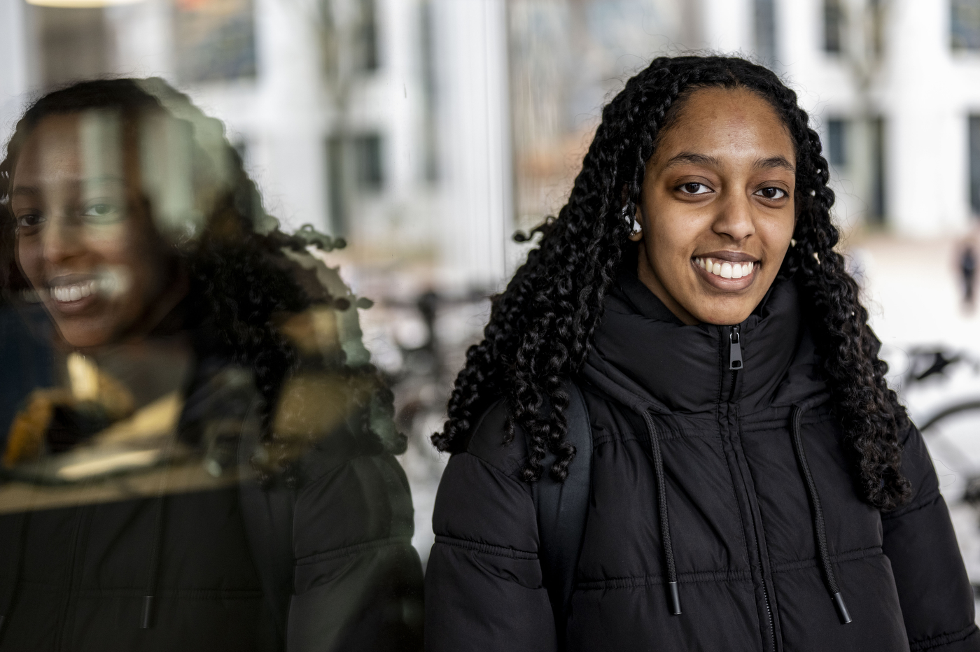 A smiling young woman with long braided hair wearing a black puffer jacket stands outdoors, with her reflection visible in a window beside her and buildings in the background.
