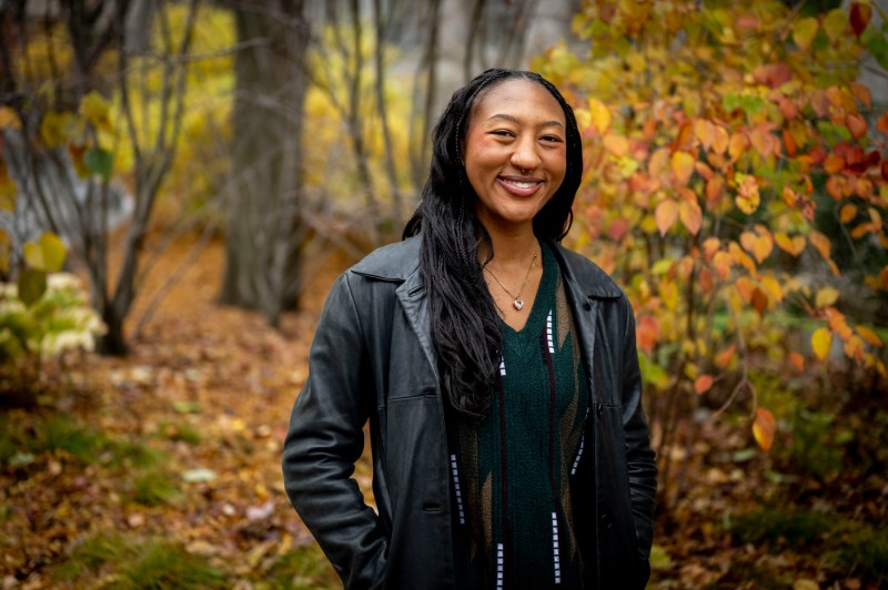 A person wearing a jacket poses for a photo outside among the foliage with their hands in their pockets.