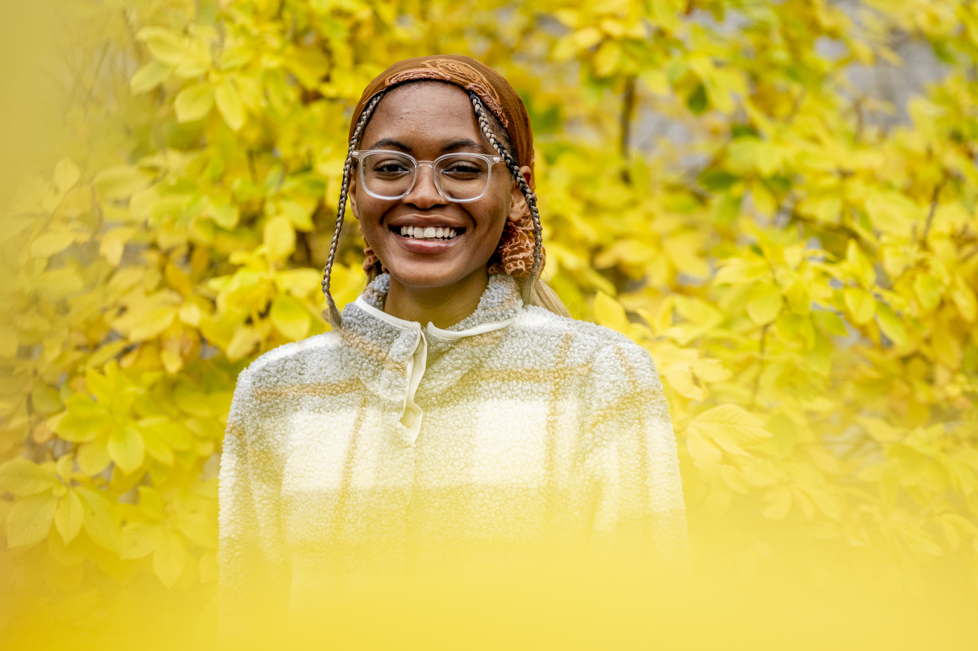 A smiling young woman wearing glasses, a patterned headscarf, and a white and yellow patterned jacket stands in front of vibrant yellow fall foliage.