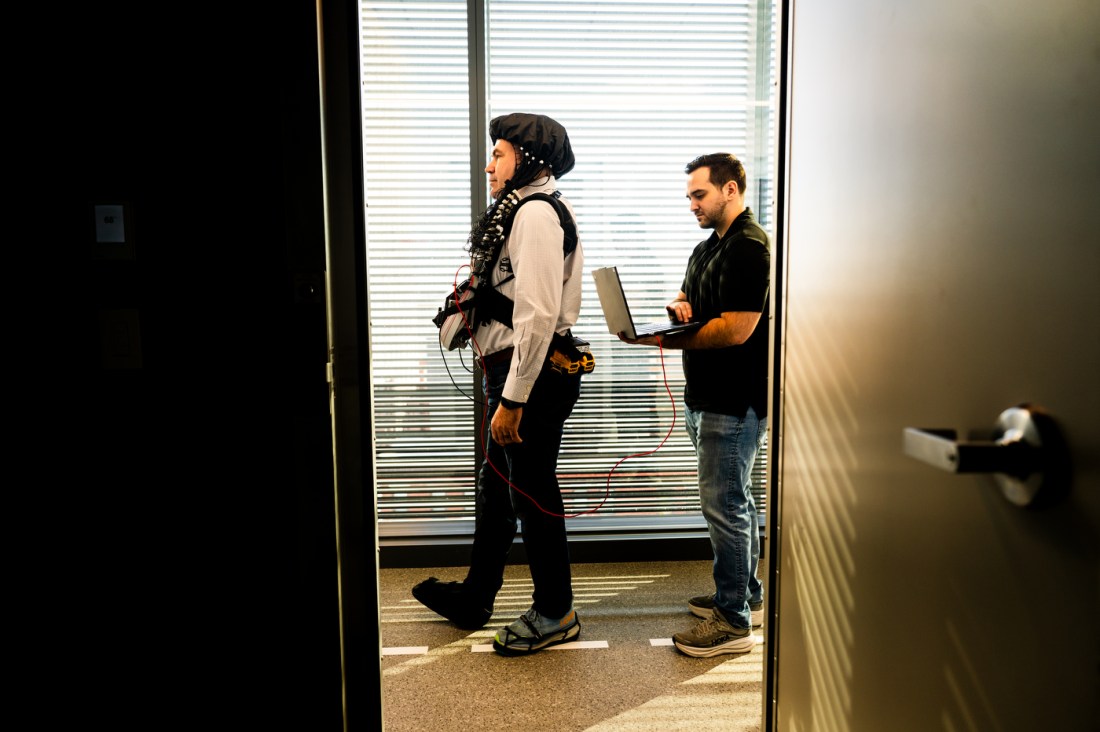 A professor wears a cap connected with wires to a vest as he walks in a lab. Another researcher follows behind him while carrying a laptop.