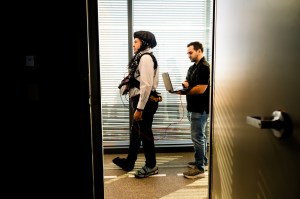 A professor wears a cap connected with wires to a vest as he walks in a lab. Another researcher follows behind him while carrying a laptop.