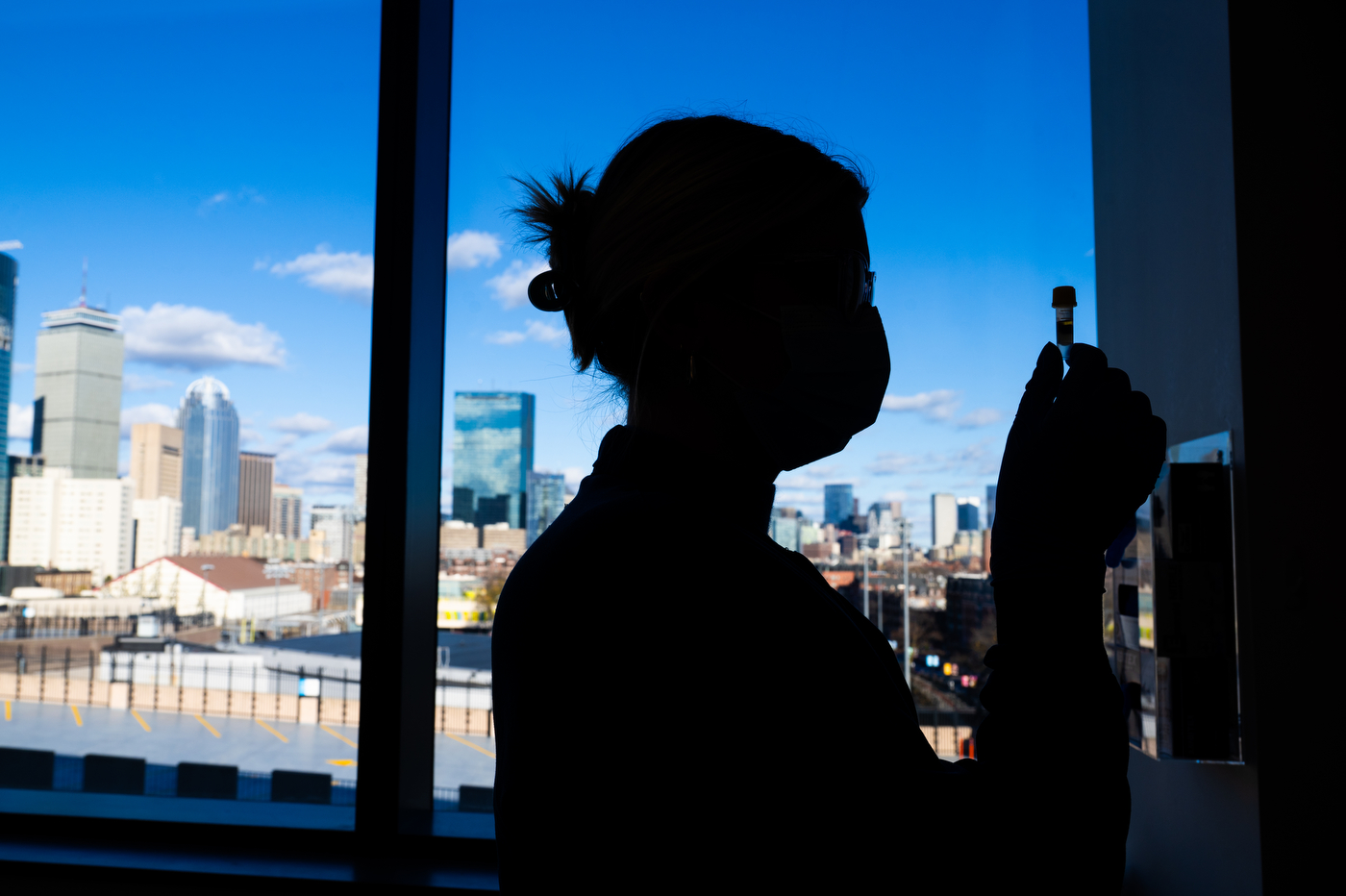 The silhouette of a person holding up a vial in front of windows, which overlook the Boston skyline.