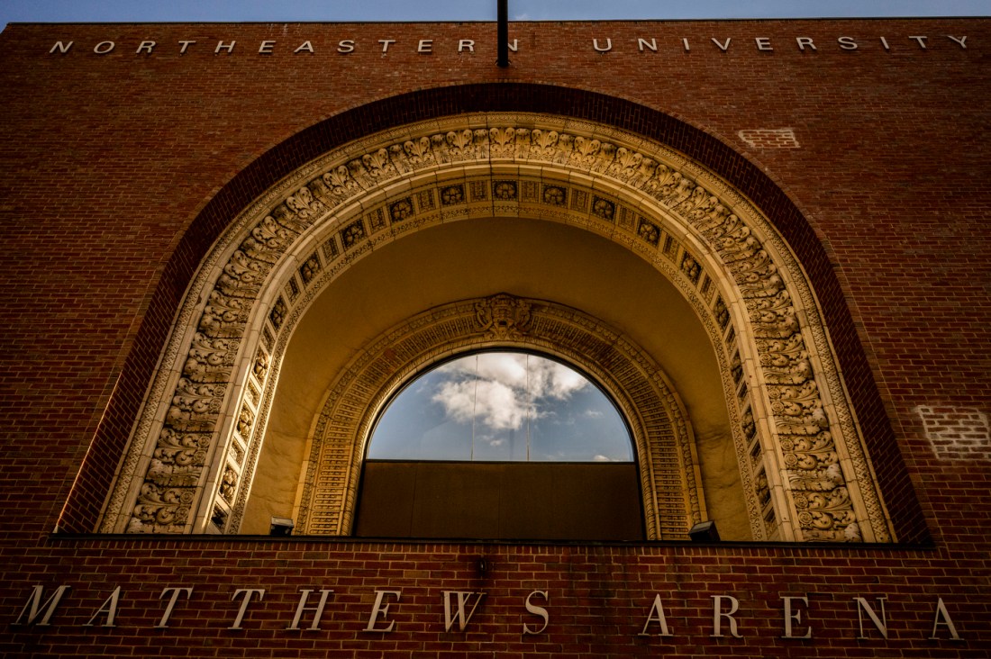 The ornate arched entrance of Matthews Arena, featuring elaborate terra cotta detailing with decorative moldings and a semi-circular window framing blue sky, set within a red brick building.
