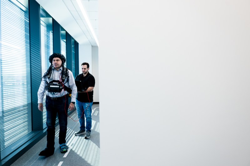 A professor walks down a hallway wearing an electrode cap as another person follows behind. 