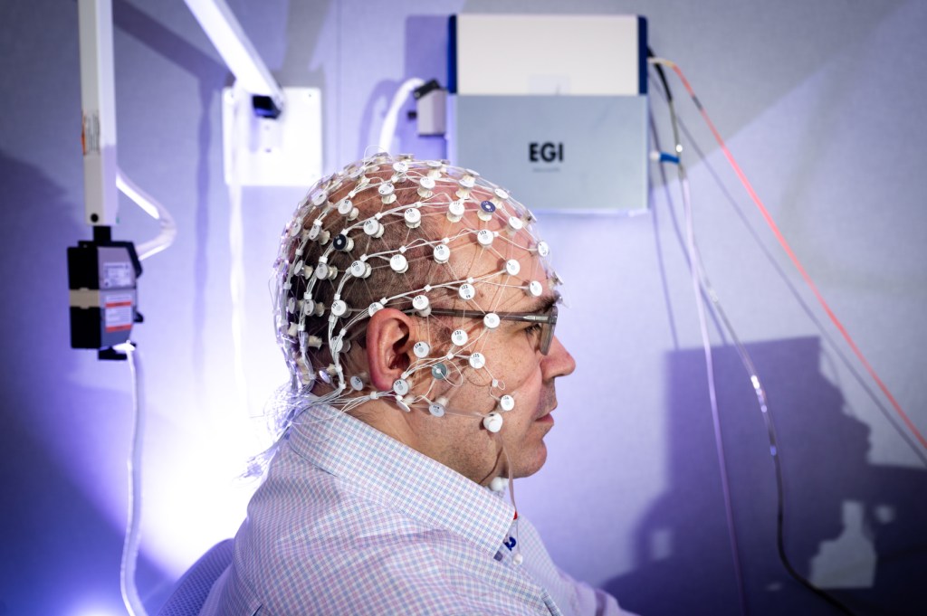 A bold man in a light purple shirt and glasses seated in profile wears an EEG electrode cap — dozens of numbered small plastic white rounds connected to each other with a net of white wires.