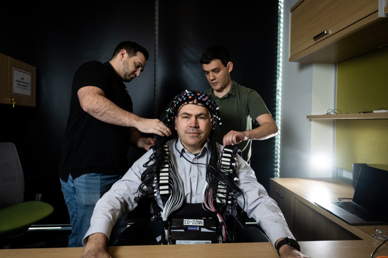 Two people hook up the cords of an electrode cap that a professor is wearing. 