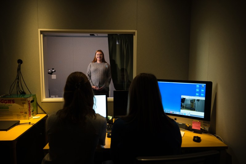 Two people tak notes on computers as a person speaks into a two-way mirror. 