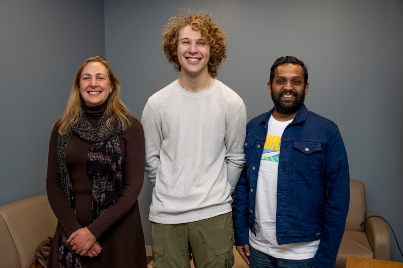Leanne Chukoskiel, William Coleman, and Sundararaman Rengarajan stand in a line and pose for a photo in front of a brown couch and grey wall. 