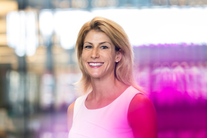 Elizabeth Glowacki smiling, wearing a pink top, standing indoors against a blurred backdrop of colorful lights in pink and purple tones.