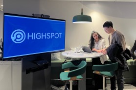 A man and a woman leaning over a laptop on a small white table, with a flatscreen displaying the text "Highspot."