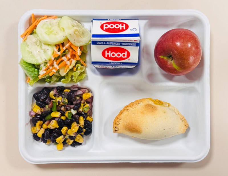A school lunch tray with compartments containing a green salad with cucumbers and shredded carrots, a carton of Hood milk, an apple, a black bean and corn salad, and an empanada or hand pie