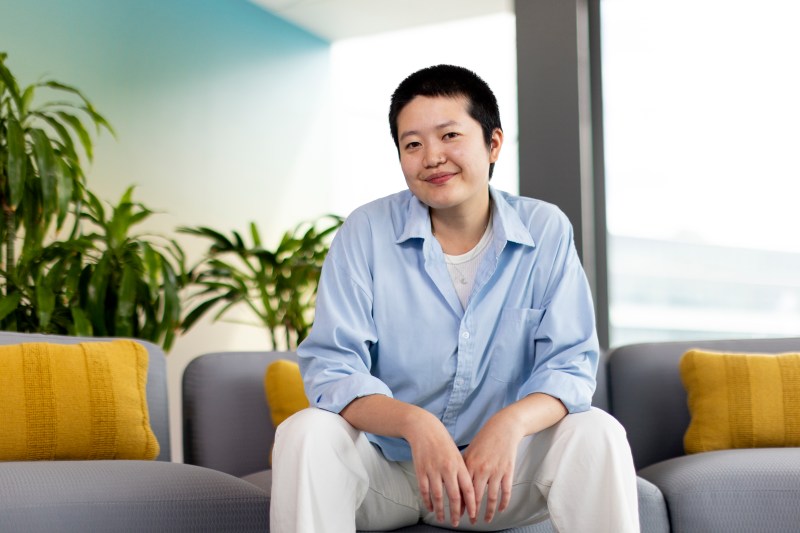 A student dressed in a blue collared shirt sits on a gray couch, smiling against a backdrop of plants.