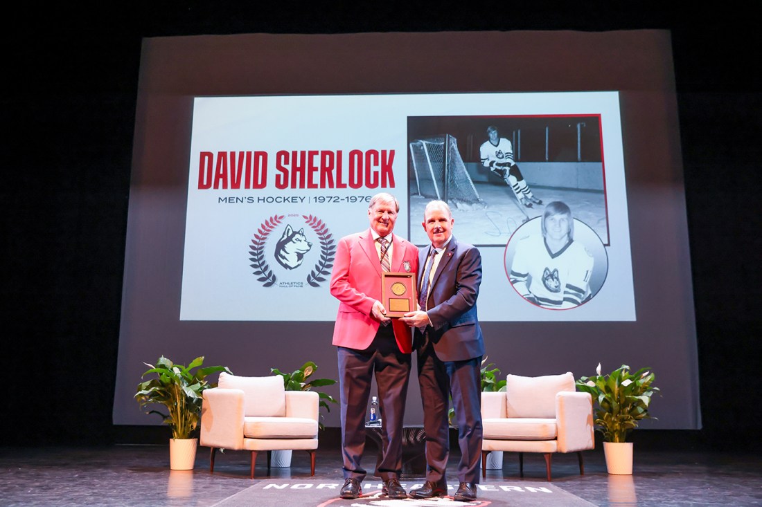 A man in a red jacket and a man in a blue jacket stand on a stage and hold a plaque in front of a projection with "David Sherlock," the Northeastern Husky logo and black and white hockey photos. 