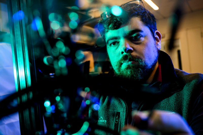 A bearded man in a dark hoodie gazes directly at the camera in a portrait lit with teal and blue lighting.