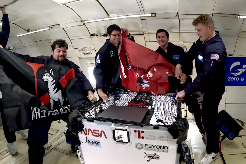 Four people in flight suits float inside a zero-gravity aircraft, holding flags and smiling. 