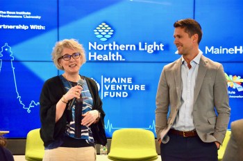 Two people stand on a stage in conversation during a presentation, with bright blue branded backdrop displaying Northern Light Health and Maine Venture Fund logos, and yellow chairs visible in the foreground.