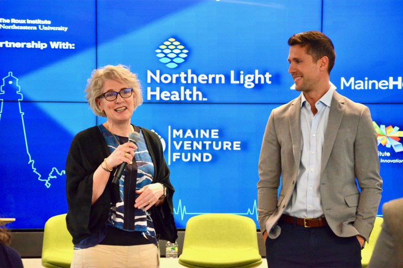 Two people stand on a stage in conversation during a presentation, with bright blue branded backdrop displaying Northern Light Health and Maine Venture Fund logos, and yellow chairs visible in the foreground.