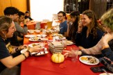 A group of male and female students eat a meal together, sitting at a long table, decorated with a red cloth, small pumpkins and dry leaves.