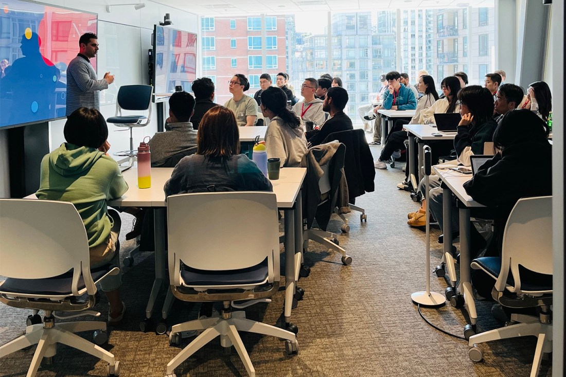 A man stands in front of a classroom full of students, lecturing in front of a large flatscreen.