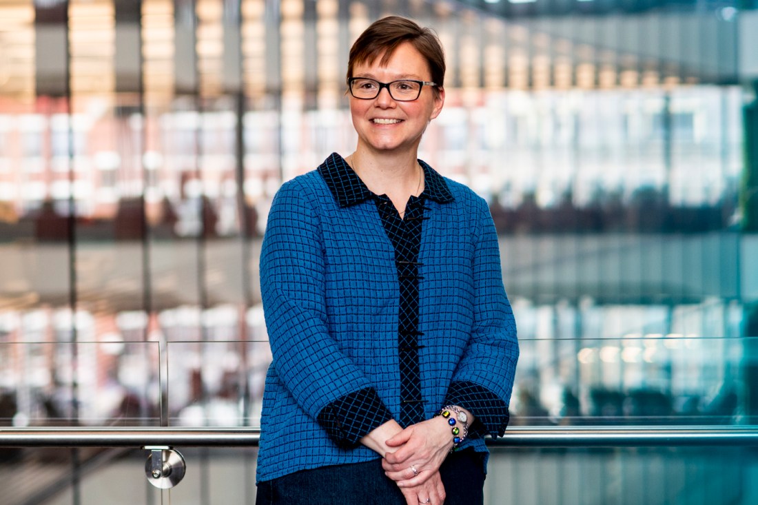 A woman with short, straight reddish hair, wearing glasses and a blouse with blue-and-black window pane fabric pattern, smiles in a half-length portrait while looking off-camera.