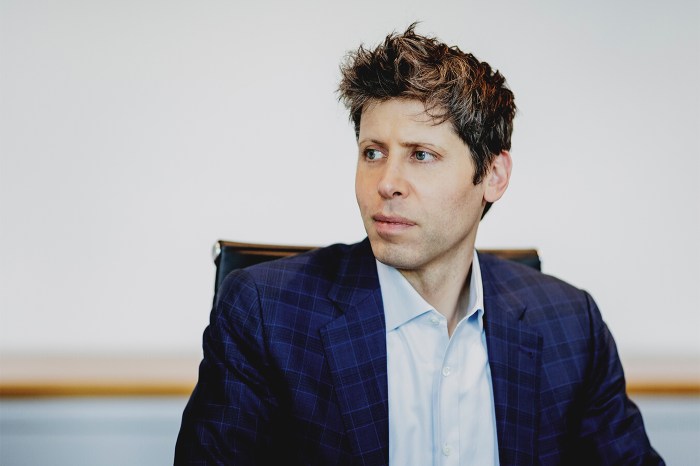 Person in a blue checkered suit jacket and light blue shirt sitting in a chair, looking to the side.