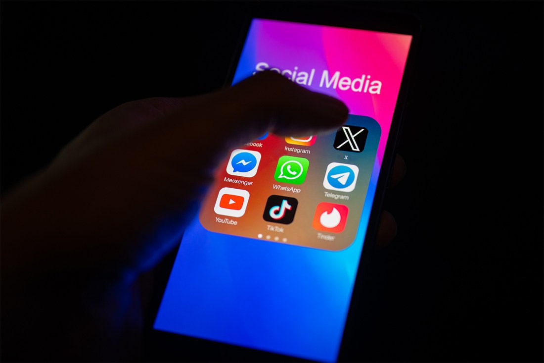 A close-up of a thumb pointing to the X/Twitter mobile app on a smartphone screen, which is displayed alongside other apps including Instagram, Telegram, Facebook, TikTok, YouTube, and Messenger.
