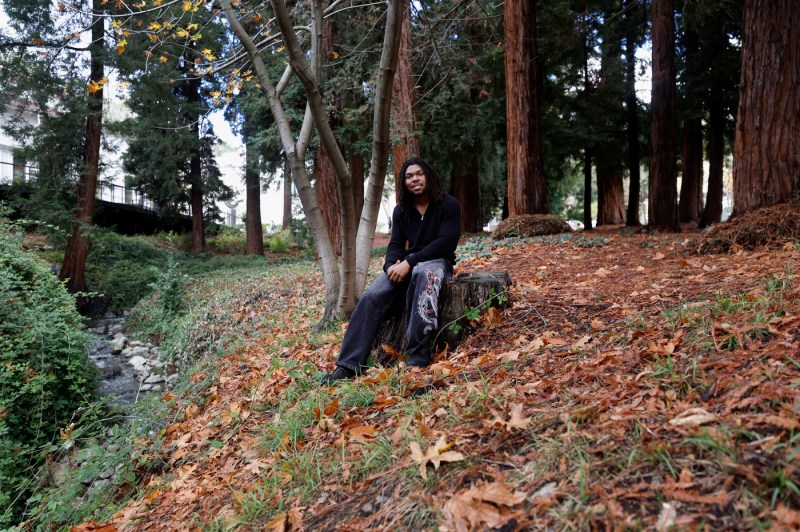 Tony Galloway is wearing a black shirt and dark blue jeans. He is sitting on a tree stump amid a pile of leaves.