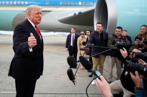 President Donald Trump gesturing to a group of reporters in front of a teal-colored jet engine.