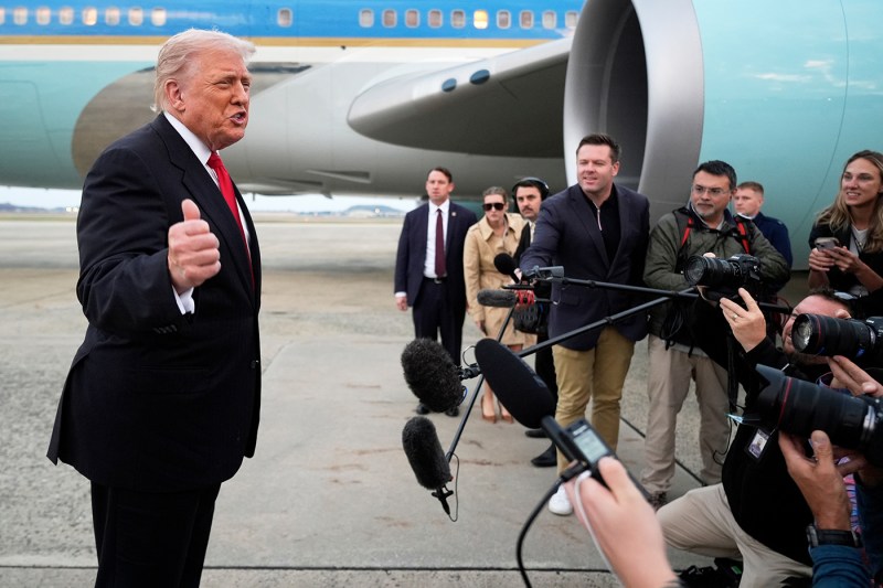 President Donald Trump gesturing to a group of reporters in front of a teal-colored jet engine.