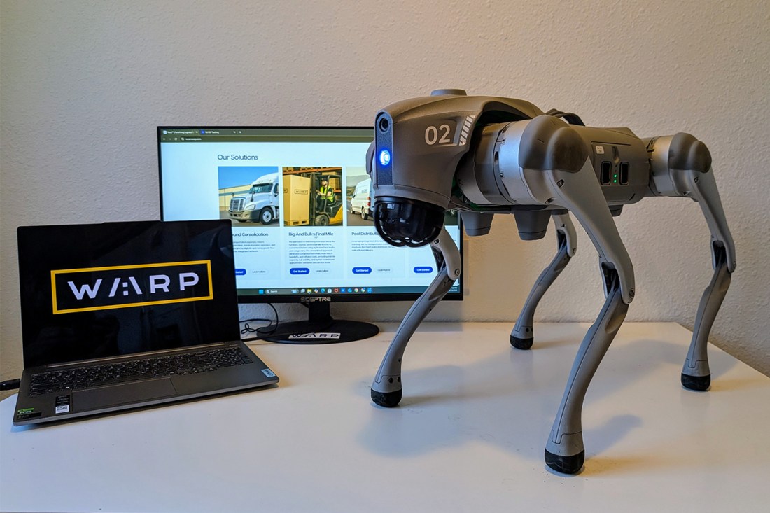 A quadruped robot dog stands on  its side on a white table next to a laptop displaying 'WARP' on its screen and a monitor showing a company website. 