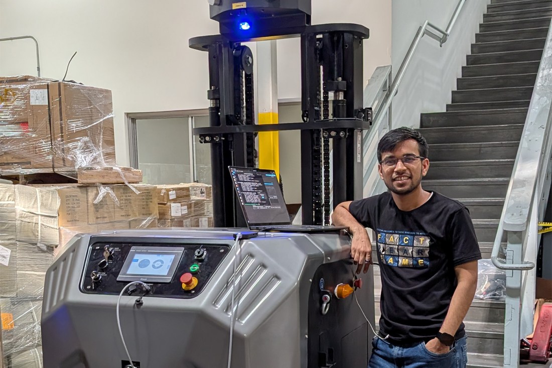 A man in a black t-shirt and jeans stands next to an industrial automated lifting machine in a warehouse setting